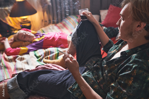 Young man smoking a cigarette and drinking wine sitting on the bed with his girlfriend relaxing near by him at home