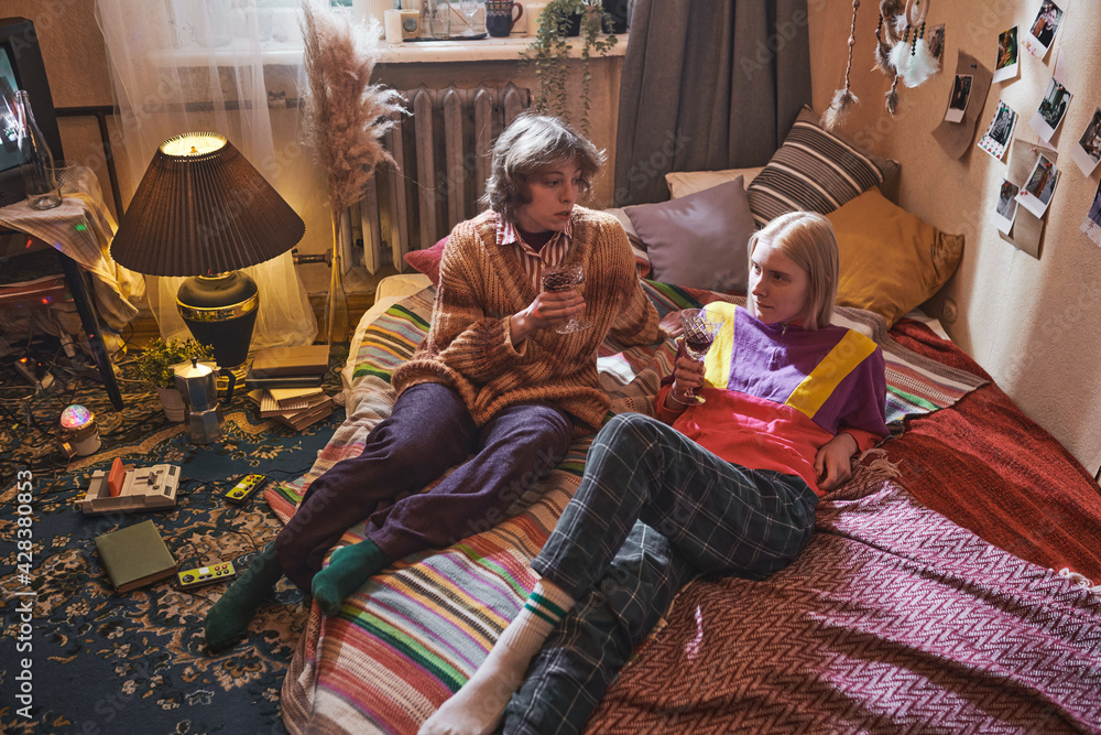 Two young friends lying on bed talking to each other and drinking red wine in the room