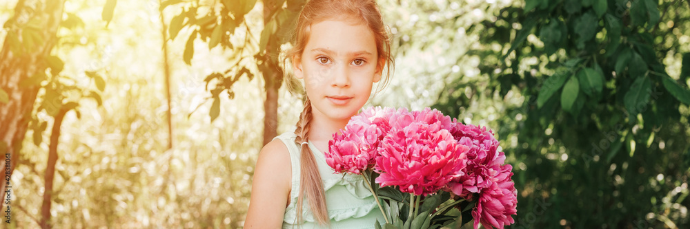 Naklejka premium portrait of a happy cute little caucasian seven year old kid girl in light green sundress, holds in hands a bouquet of pink peony flowers in full bloom on the background of nature. banner. flare