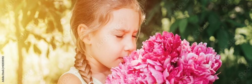 portrait of a happy cute little caucasian seven year old kid girl, holds in hands and smell and enjoying a bouquet of pink peony flowers in full bloom on the background of nature. banner. flare portrait of a happy cute little caucasian seven year old kid girl, holds in hands and smell and enjoying a bouquet of pink peony flowers in full bloom on the background of nature. banner. flare