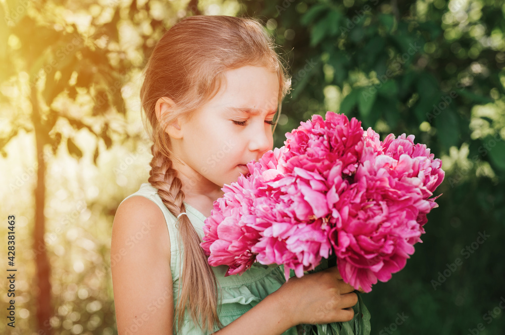 Fototapeta premium portrait of a happy cute little caucasian seven year old kid girl, holds in hands and smell and enjoying a bouquet of pink peony flowers in full bloom on the background of nature. flare