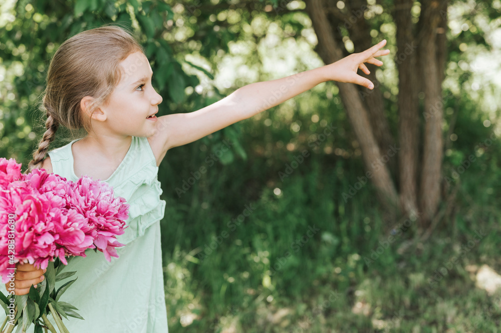portrait of a happy cute little caucasian seven year old kid girl, holds in hands a bouquet of pink peony flowers in full bloom and pointing to the side on the background of nature portrait of a happy cute little caucasian seven year old kid girl, holds in hands a bouquet of pink peony flowers in full bloom and pointing to the side on the background of nature