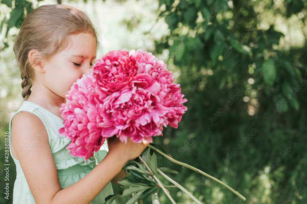 Naklejka premium portrait of a happy cute little caucasian seven year old kid girl, holds in hands and smell and enjoying a bouquet of pink peony flowers in full bloom on the background of nature