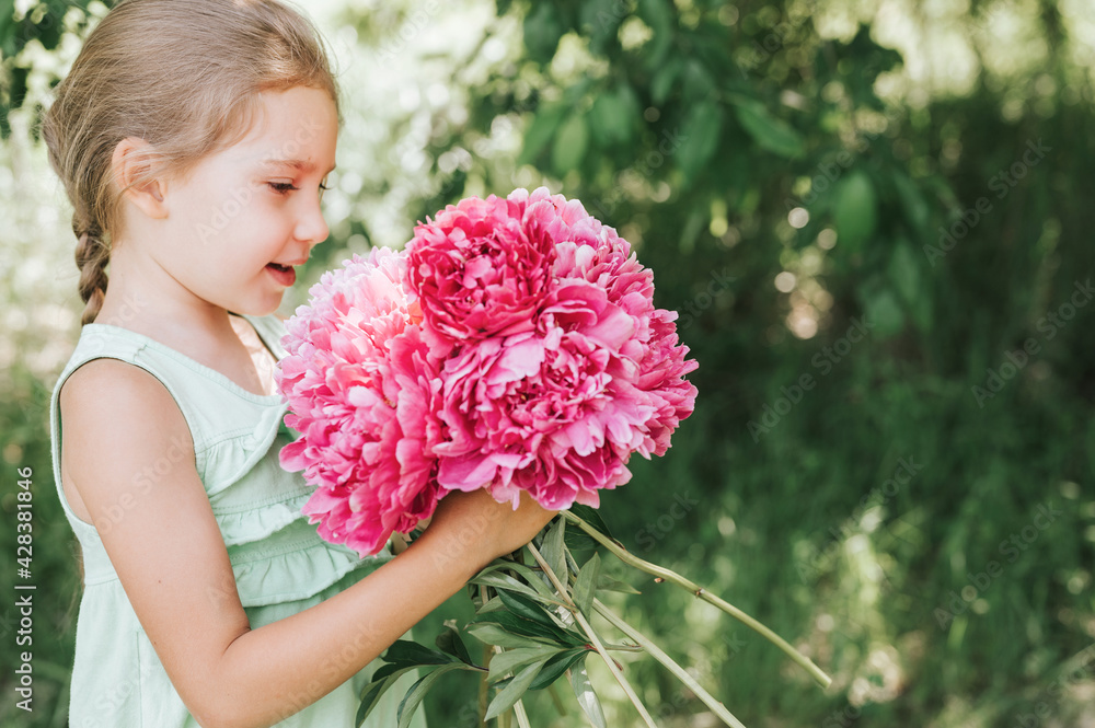 Fototapeta premium portrait of a happy cute little caucasian seven year old kid girl, holds in hands and smell and enjoying a bouquet of pink peony flowers in full bloom on the background of nature
