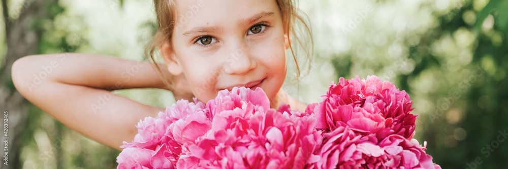 portrait of a happy cute little caucasian seven year old kid girl, holds in hands a bouquet of pink peony flowers in full bloom and straightening her hair on the background of nature. banner portrait of a happy cute little caucasian seven year old kid girl, holds in hands a bouquet of pink peony flowers in full bloom and straightening her hair on the background of nature. banner