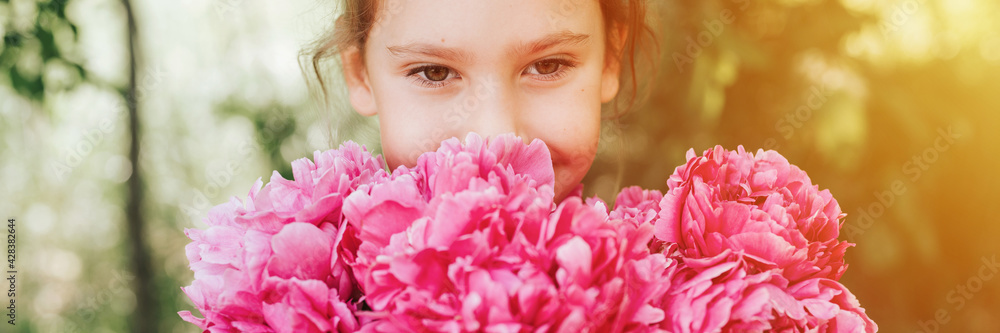 Fototapeta premium portrait of a happy cute little caucasian seven year old kid girl, holds in hands and smell and enjoying a bouquet of pink peony flowers in full bloom on the background of nature. banner. flare