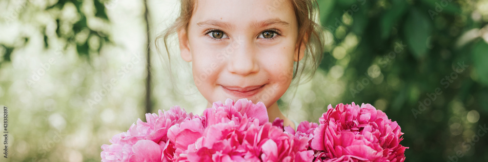 Fototapeta premium portrait of a happy cute little caucasian seven year old kid girl, holds in hands a bouquet of pink peony flowers in full bloom on the green background of nature. banner
