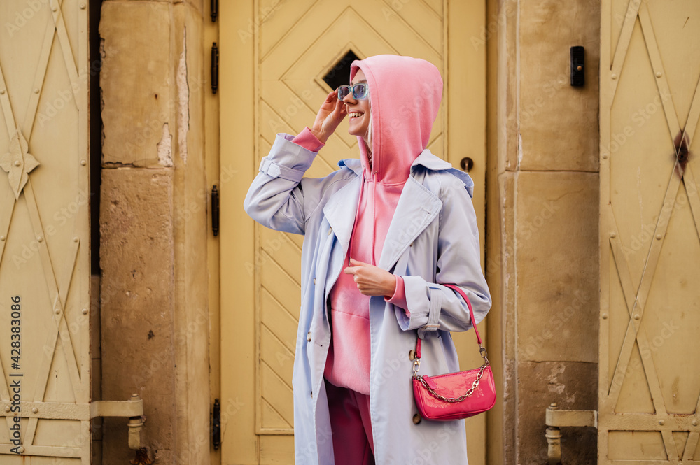 Happy smiling fashionable woman wearing trendy sport chic style outfit posing in street of European city. Blue trench coat, sunglasses, pink hoodie, mini baguette bag. Copy, empty space for text