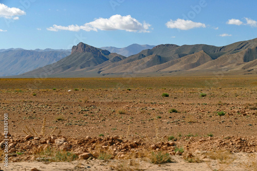 View of a stone desert field in front of beautiful brown vegetation-free rolling mountains and blue sky with little white clouds between Midelt and the Gorges du Ziz in Morocco, Africa 