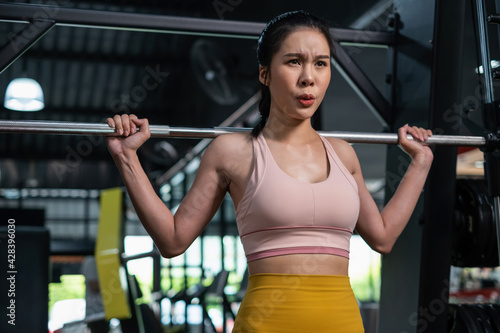 Young Asia woman lifting barbells in the fitness gym. Doing shoulder press exercise with a weight bar. Flexing muscles and making shoulder press squat. Workout and healthy lifestyle.