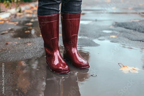 Female feet in red rubber boots stand in muddy puddle under rain in autumn weather