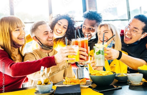 Multiracial friends wearing face masks drinking cocktails at bar restaurant - New normal friendship concept with young people having fun at home party - Focus on glasses 
