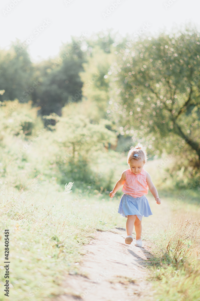 little girl 2-3 years old European appearance. the child walks on a path near the forest. blonde hair. green forest and grass. sunny day.