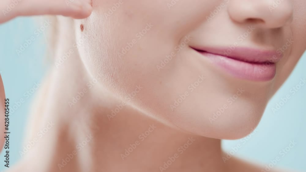 Extreme close-up of female beauty model touches her jawline smiling for the camera against blue background | Moisturizer wearing shot for face care commercial Extreme close-up of female beauty model touches her jawline smiling for the camera against blue background | Moisturizer wearing shot for face care commercial