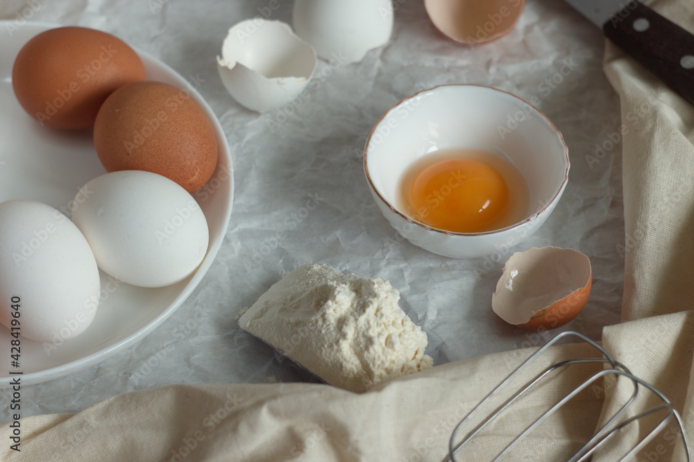 In the kitchen for cooking on the table - eggs on a plate, a broken egg with yolk in a bowl, a shell, a slide of flour and textiles In the kitchen for cooking on the table - eggs on a plate, a broken egg with yolk in a bowl, a shell, a slide of flour and textiles