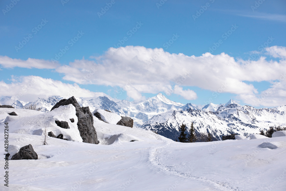 Winter hiking trail, snowcapped mountains with cloudscape, blue sky. Stones at the top of the mountain covered by snow, path in the snow. Swiss alps, Wallis canton.
