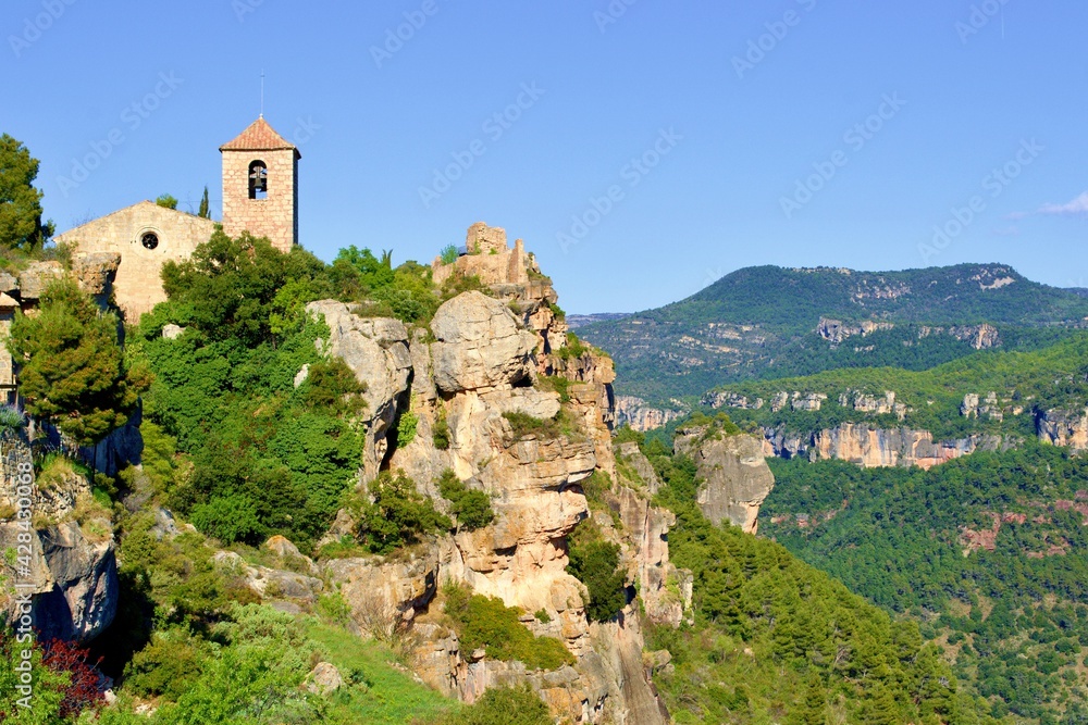 Obraz premium Romanesque church of Santa Maria de Siurana on top of rock, view into valley, mountains on the background. Blue sky with white clouds. Siurana, Catalonia, Spain.