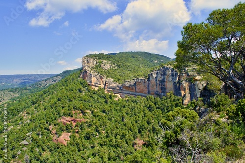 Orange massive rock on green mountainside covered with trees, climbing sector. Blue sky with white clouds. Beautiful nature, mountains. Siurana, Catalonia, Spain.