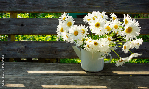 a bouquet of daisies in a white teapot on an old bench in the garden