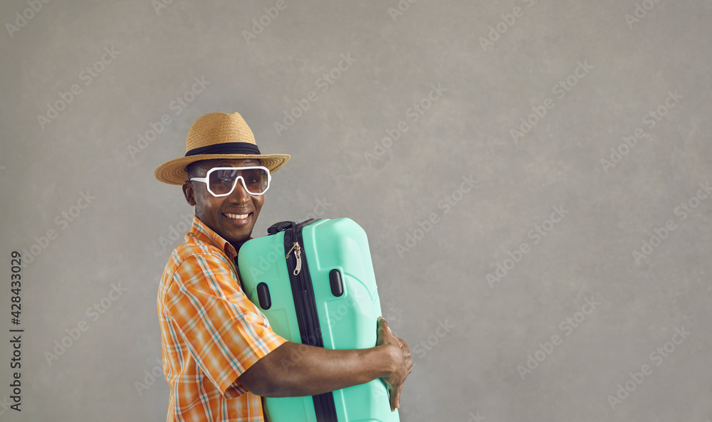 Banner with portrait of happy black tourist with his baggage. African man in sunglasses, sun hat and orange shirt smiling and holding mint green travel suitcase isolated on gray copy space background Banner with portrait of happy black tourist with his baggage. African man in sunglasses, sun hat and orange shirt smiling and holding mint green travel suitcase isolated on gray copy space background