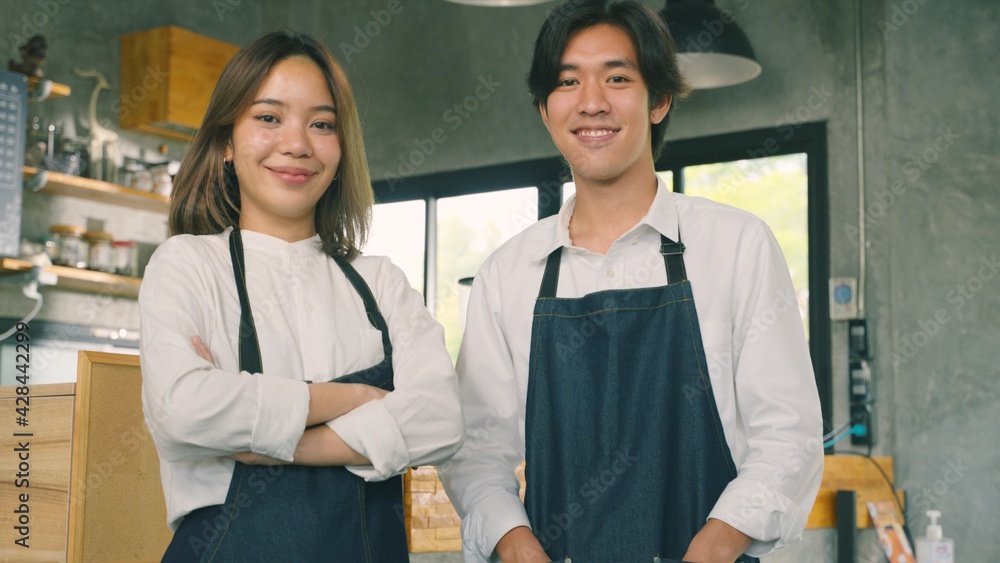 Portrait of young Asian colleague standing at their new business. Couple owner standing inside restaurant. Waitresses in aprons smiling standing together and looking at camera. Portrait of young Asian colleague standing at their new business. Couple owner standing inside restaurant. Waitresses in aprons smiling standing together and looking at camera.