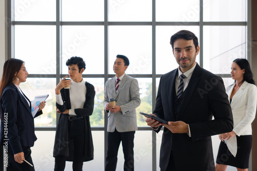 portrait of young caucasian confident businessman with smiling face stnading in office with background of multiethnic colleagues. diversity in business concept