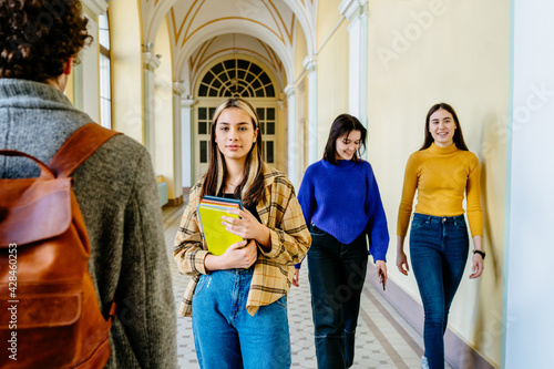 Portrait of young female student standing alone keeping book stack looking at camera with students in bright colorful clothes at the background and foreground. Education, College, school, University. photo