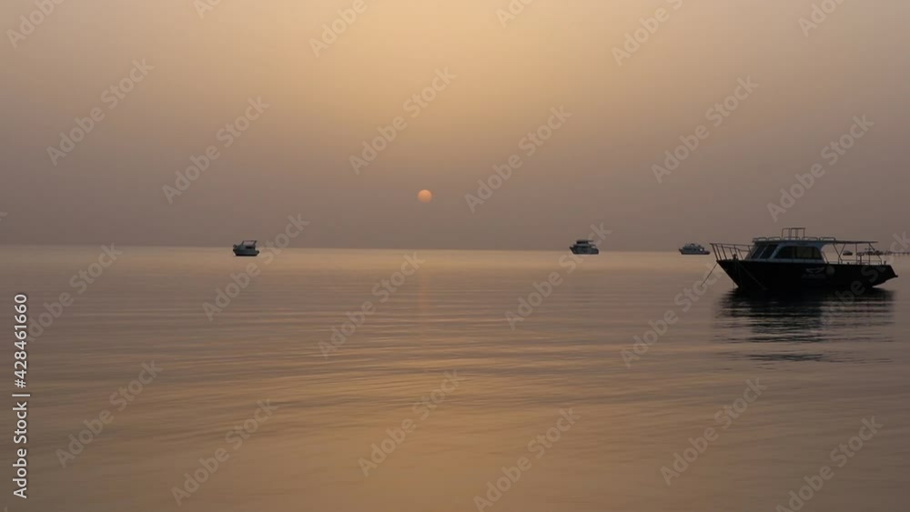Dawn on the Red Sea in Egypt, Makadi Bay, calm sea, silhouettes of boats, golden disc of the sun on the horizon, reflection of sunlight on the water surface Dawn on the Red Sea in Egypt, Makadi Bay, calm sea, silhouettes of boats, golden disc of the sun on the horizon, reflection of sunlight on the water surface