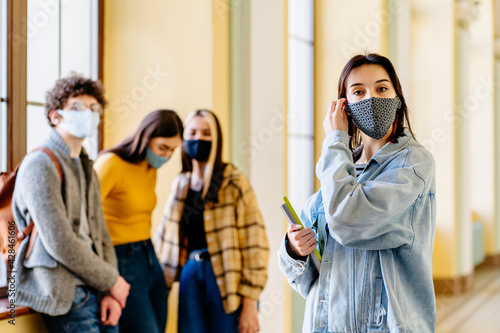Portrait of young white girl student wearing protective mask wearing jeans jacket and striped shirt posing in university hall with other students on blurred background and pile or books.
