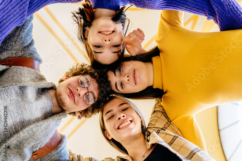 Group of young students standing in a circle, laughing, hugging indoors, having fun, hands Several people of different nationalities smiling and looking down at camera. Concepts friendship, teamwork.