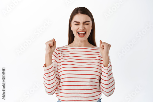 Hooray I did it. Excited successful girl celebrating achievement, winning and shouting yes, making fist pump, gain her goal, triumphing of victory, standing over white background