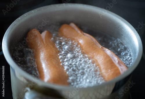 Two sausages are boiling in water, inside a metal bowl. Close up.
