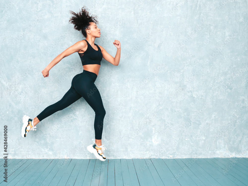 Fitness smiling black woman in sports clothing with afro curls hairstyle.She wearing sportswear. Young beautiful model with perfect tanned body.Female jumping and running in studio near gray wall