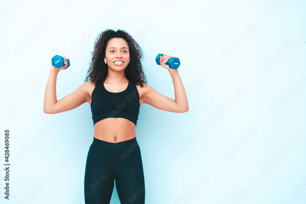 Fitness smiling black woman in sports clothing with afro curls hairstyle.She wearing sportswear. Young beautiful model with perfect tanned body.Female holding dumbbells in studio near light blue wall Fitness smiling black woman in sports clothing with afro curls hairstyle.She wearing sportswear. Young beautiful model with perfect tanned body.Female holding dumbbells in studio near light blue wall