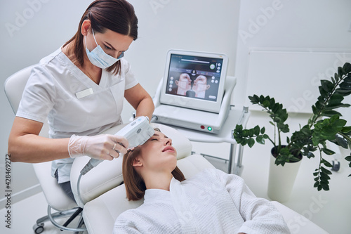 Charming Caucasian redheaded woman in white bathrobe receiving cleaning skin face with ultrasound modern device in beauty center photo