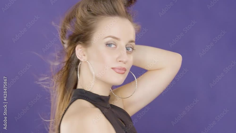Charming beautiful cute young girl brunette model with beautiful eyes poses for camera and touches wind blowing hair on purple background in foto studio Charming beautiful cute young girl brunette model with beautiful eyes poses for camera and touches wind blowing hair on purple background in foto studio