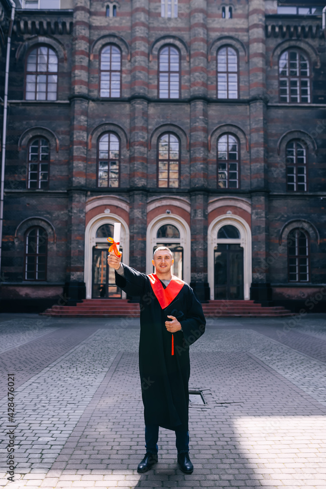 a handsome, tall man after graduation stands on the street in front of the university building holding a diploma rewound with a red ribbon.