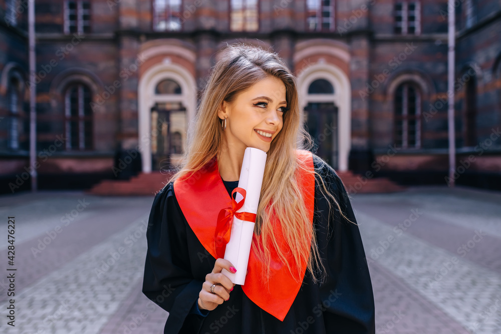 Fototapeta premium A young girl with long hair, a graduate holding a diploma on the background of the university.