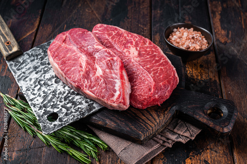 Uncooked Raw Shoulder Top Blade or flat iron beef meat steaks on a wooden butcher board with meat cleaver. Dark wooden background. Top View