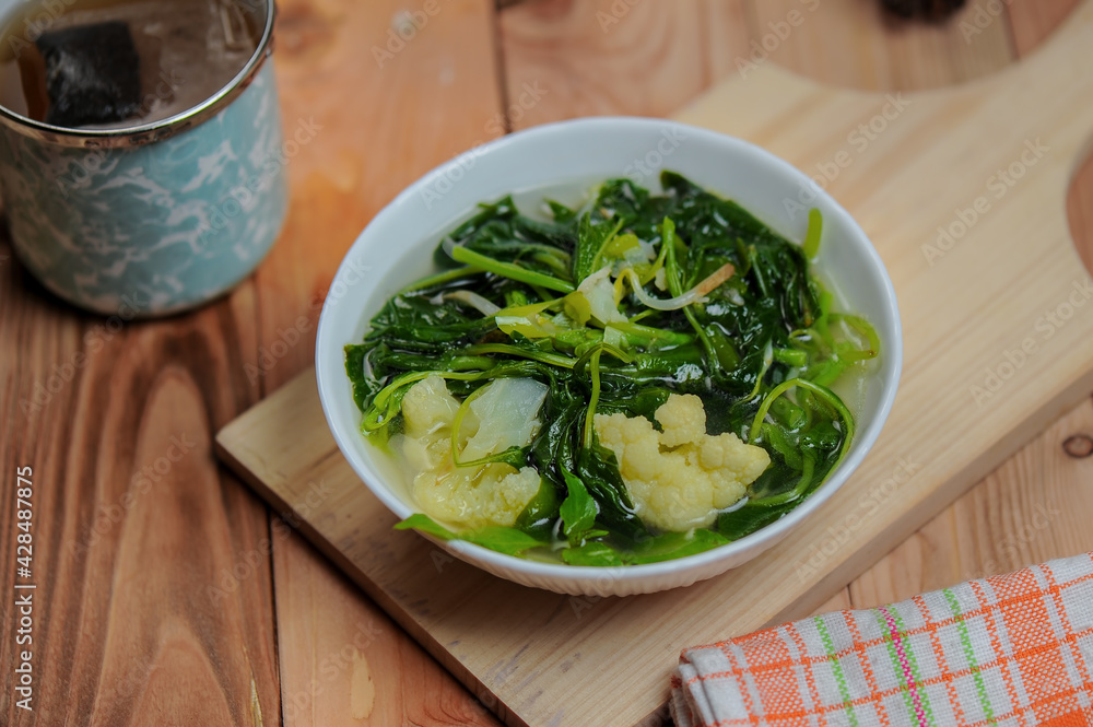 Sayur Bening Bayam or spinach clear soup with broccoli in a white bowl and wooden table Sayur Bening Bayam or spinach clear soup with broccoli in a white bowl and wooden table