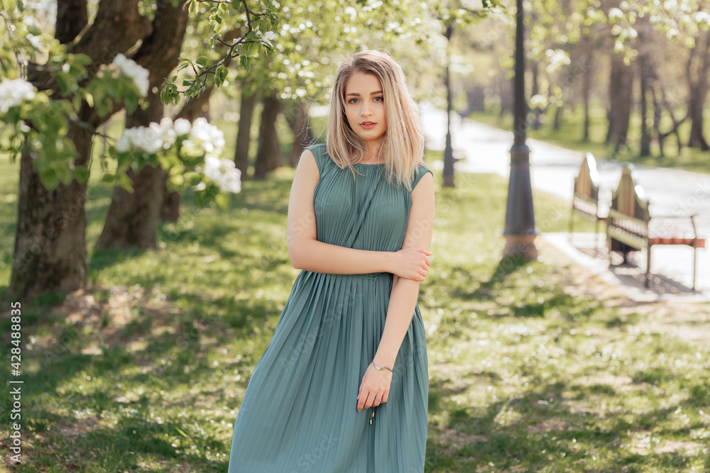 Beautiful blonde woman in a green long dress stays in a spring blooming garden. Beautiful blonde woman in a green long dress stays in a spring blooming garden.