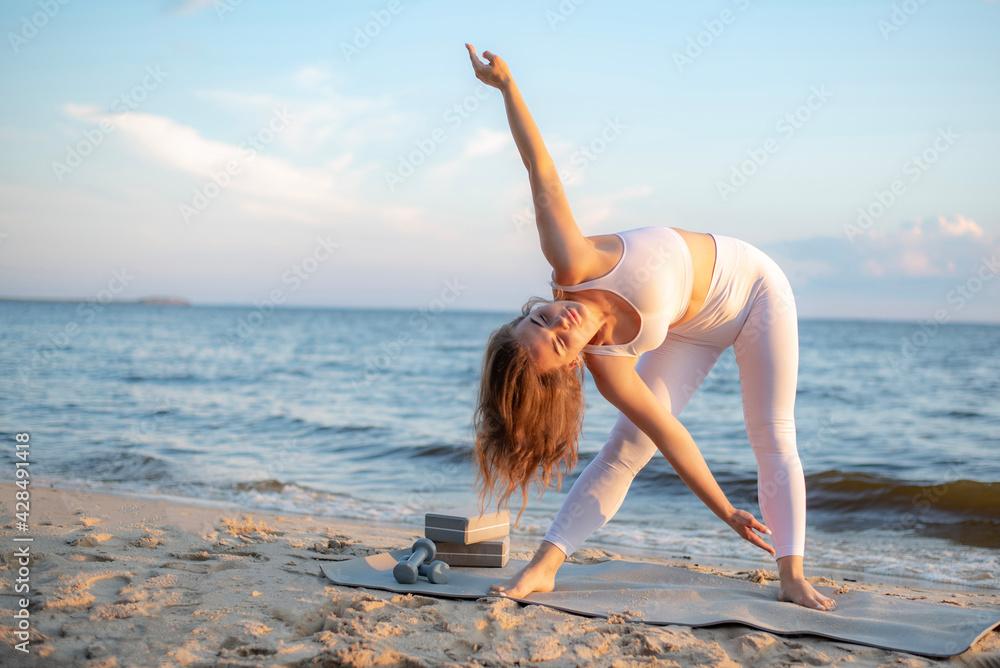 athletic girl doing hand warm-up before yoga class. charming young woman in a blue sports tight-fitting suit on the coast enjoying the sunset. sports girl is engaged in stretching on a yoga mat. athletic girl doing hand warm-up before yoga class. charming young woman in a blue sports tight-fitting suit on the coast enjoying the sunset. sports girl is engaged in stretching on a yoga mat.