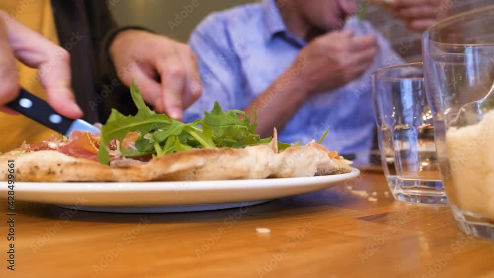 Time-lapse fast motion side view of adult and senior man - in background eating delicious pizza made from organic bio ingredients with fresh rucola arugula and prosciutto in traditional restaurant piz Time-lapse fast motion side view of adult and senior man - in background eating delicious pizza made from organic bio ingredients with fresh rucola arugula and prosciutto in traditional restaurant piz