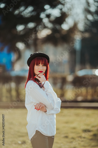 Vertical side view of a young smiling female with red hair, sunglasses, and a hat posing in a park photo