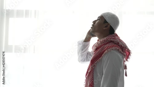 portrait of muslim man using hand to pray against white window. takbir and adzan