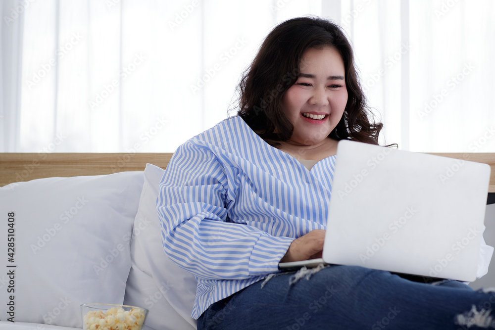 Asian young happy fat chubby woman lay down have fun laughing happily lean on white pillow on bed using laptop computer to watch movie and eat popcorn on quarantine lockdown on covid pandemic at home