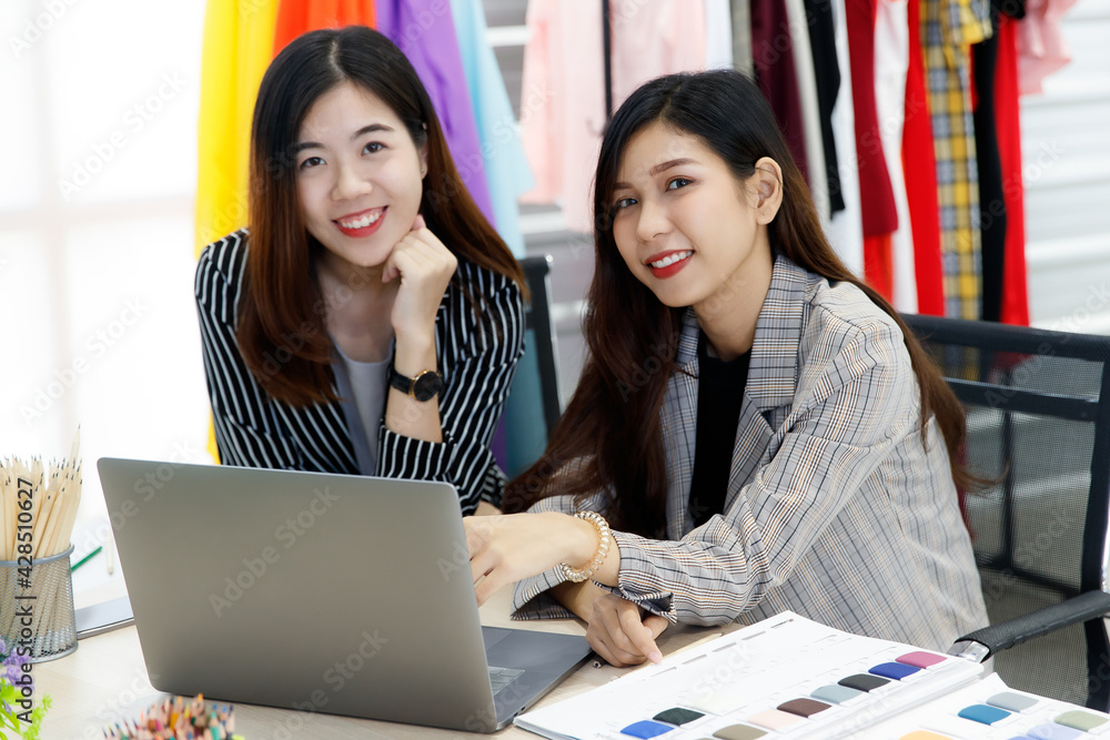 Two Asian young happy successful female stylish fashion designer wears casual suit sit smiling look at camera when using laptop computer working in front cloth rack in creative design studio workshop Two Asian young happy successful female stylish fashion designer wears casual suit sit smiling look at camera when using laptop computer working in front cloth rack in creative design studio workshop