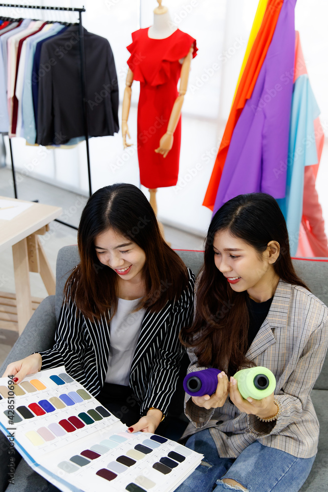 Fototapeta premium Two Asian young happy successful beautiful female creative stylish fashion designer colleagues hold fabric color pattern book and thread roll in hands talking discussing together on sofa in studio
