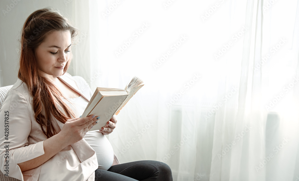 Young pregnant woman reading a book while sitting on the sofa at home. Young pregnant woman reading a book while sitting on the sofa at home.