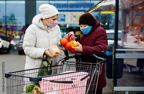 Senior woman and social worker with medical mask due pandemic coronavirus disease. Daughter or granddaughter help grandmother with shopping in supermarket, push cart trolley with foods, outdoors photo